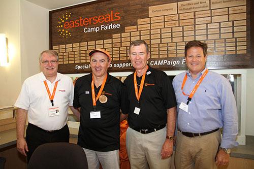 Four men stand side by side in front of a large donor wall at Camp Fairlee. They are all wearing orange lanyards and smiling at the camera.