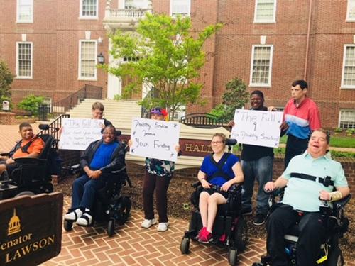 A group of people with disabilities gather outside of legislative hall with signs