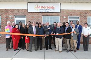 A group of people stand in front of Easterseals Smyrna. They are all smiling at the camera as a man in the center cuts a ribbon with large scissors.