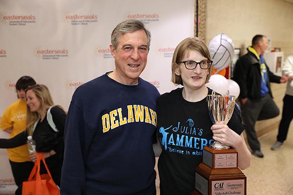 Person with disability holds a trophy and poses with elected official at volleyball event