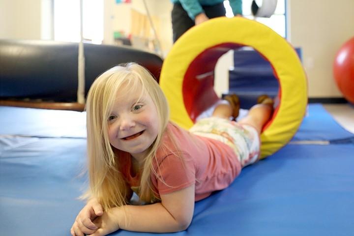 A smiling child with blonde hair lays on her stomach in a colorful play room