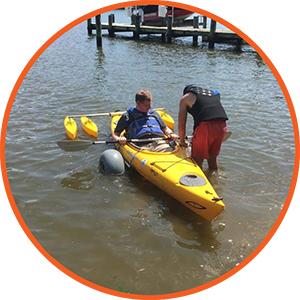 A camper in an adaptive kayak. To their right, a counselor is bending over to assist them.