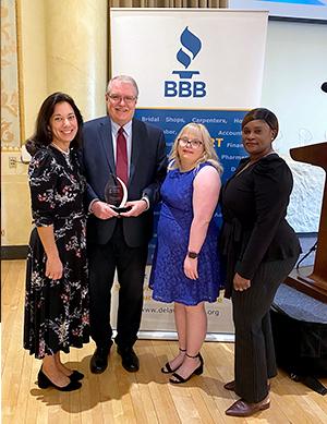 A group of individuals stand in front of a BBB backdrop. The man in the center is holding an award.