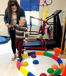 A child with a disability walks along a circular beam as a therapist assists him.