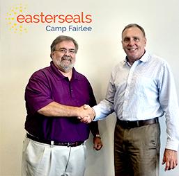 Two men shake hands and smile at camera while standing in front of an Easterseals Camp Fairlee sign