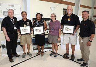 A group stands side by side posing for the camera. They are each holding a drawing of Camp Fairlee.