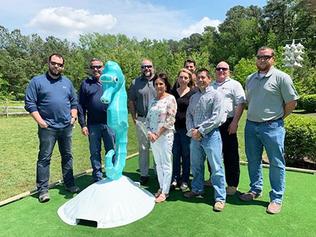 A group of people stand on a gold course next to a sculpture of a blue seahorse.