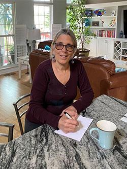 Woman sitting at desk writing on a piece of paper while she looks at the camera