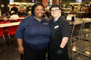 A man with Down Syndrome stands next to a support professional in a cafe