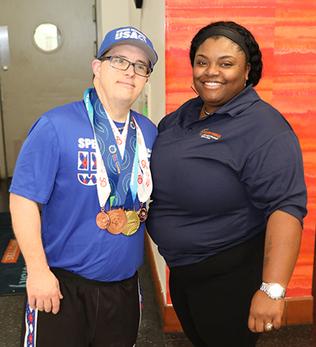 Young adult male with Down syndrome wearing a hat and several special Olympic medals standing next to he support specialist who is female with short black hair