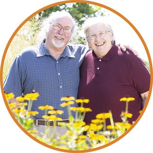 Two elderly people stand in a garden smiling at the camera