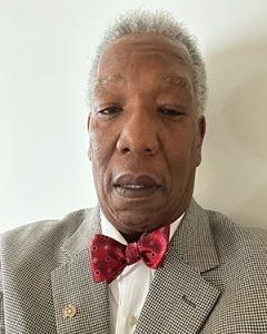 A headshot of a man with a red bowtie and beige suit looking at the camera.