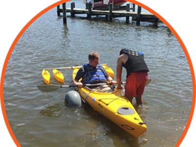 A camper in an adaptive kayak. To their right, a counselor is bending over to assist them.