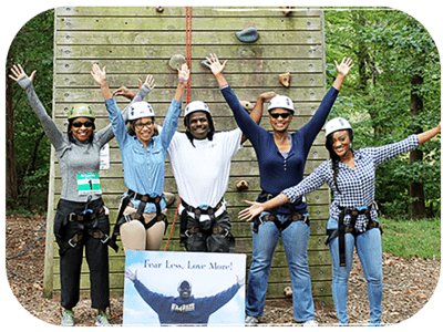 A group of people wearing helmets standing in front of a climbing tower at camp. Their arms are stretched out above their heads and they are standing in front of a memorial sign.