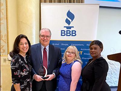 A group of individuals stand in front of a BBB backdrop. The man in the center is holding an award.