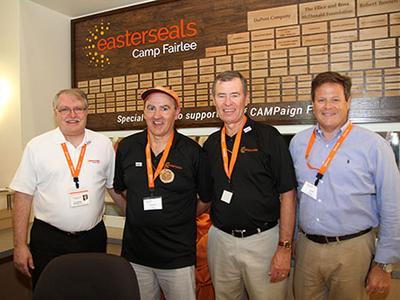 Four men stand side by side in front of a large donor wall at Camp Fairlee. They are all wearing orange lanyards and smiling at the camera.