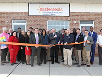 A group of people stand in front of Easterseals Smyrna. They are all smiling at the camera as a man in the center cuts a ribbon with large scissors.