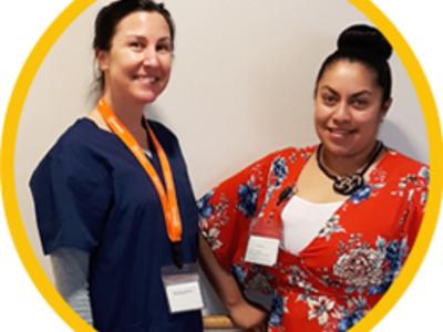 A nurse and an assistant wearing scrubs smile at the camera