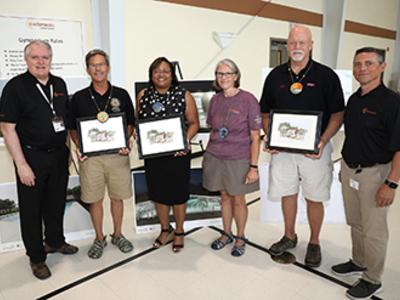 A group stands side by side posing for the camera. They are each holding a drawing of Camp Fairlee.