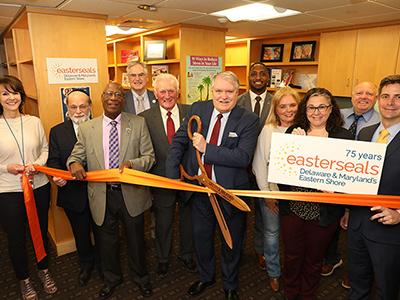 A group of people stand in a resource center. The man in the center is cutting a ribbon with large scissors.