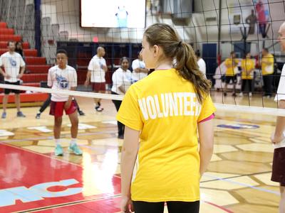 A woman with a ponytail and a yellow volunteer shirt stands on a volleyball court