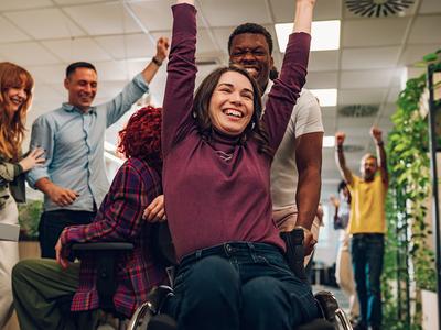 A woman in a wheel chair smiles and cheers while a room behind her does the same