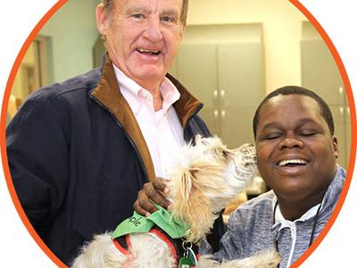 Volunteer holds up a dog that licks the face of a participant. The volunteer is smiling to the camera.