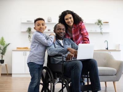 A man in a wheelchair and a laptop on his lap smiles at the camera with his wife and child by his side.