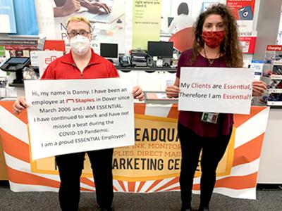 Two individuals wearing masks, stand in front for a bright colored table, while they hold signs