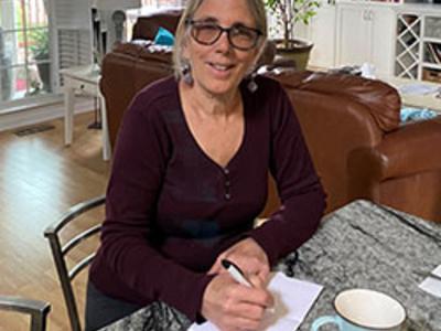 Woman sitting at desk writing on a piece of paper while she looks at the camera