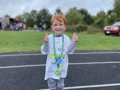 young child with red hair with race medal around his neck giving thumbs up