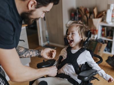 A man is helping strap a child into a wheelchair while the child is laughing