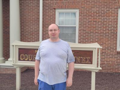 Adult male in a grey shirt standing infront of his place of employment