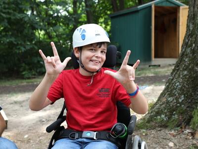 Boy with a disablity at camp in a wheelchair smiling as they stroll through the wooded trail