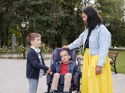 A child in a wheel chair in a park is accompanied by a woman and young boy