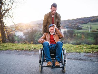 A man is sitting in a wheelchair as his son is behind him pushing him in a scenic rural setting