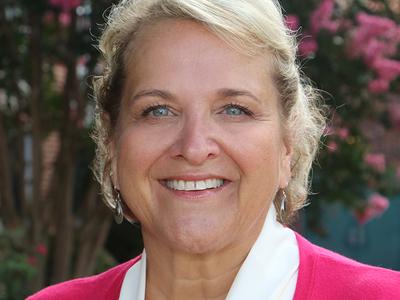 A headshot of a woman with blonde hair standing in front of a pink bush. She is wearing bright pink and is smiling.