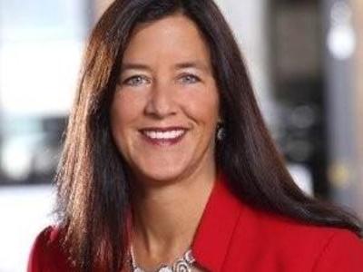 A headshot of a woman with long brown hair, wearing a red jacket and smiling at the camera