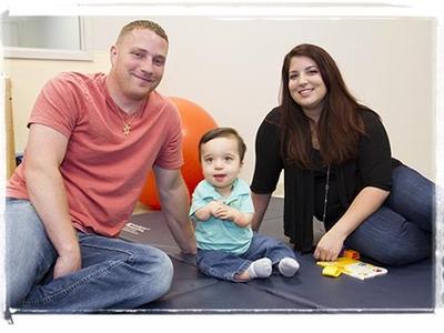 In a play area, a child sits on a floor mat while his parents sit to either side of him. They are all smiling at the camera