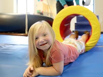 A smiling child with blonde hair lays on her stomach in a colorful play room