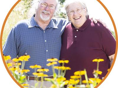 Two elderly people stand in a garden smiling at the camera