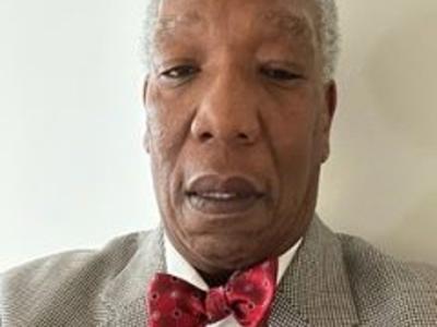 A headshot of a man with a red bowtie and beige suit looking at the camera.