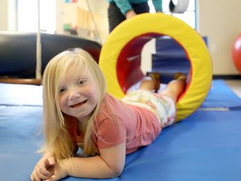 A child lays on a mat in a sensory gym and smiles to the camera.