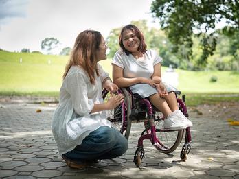 Two women are speaking to each other. One woman is in a wheel chair and the other woman is kneeling beside her.