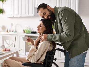 A woman in a wheel chair is seated for breakfast and is comforted by her husband who stands behind her.