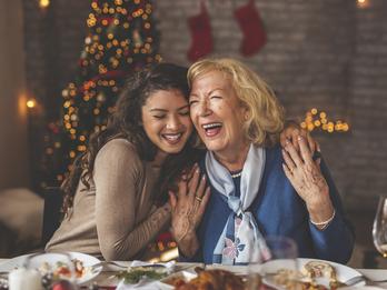 A mother and daughter are at a dining room table eating dinner and smiling together. In the background are holiday decorations.