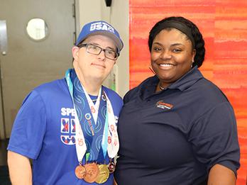 Young adult male with Down syndrome wearing a hat and several special Olympic medals standing next to he support specialist who is female with short black hair